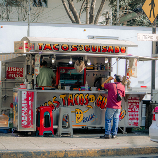 A bustling street vendor selling tacos in a colorful open-air stall on a sunny day.